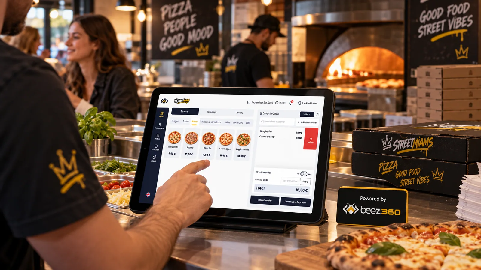 Order taking on BeezPOS in front of a pizza oven, with a Beez360 sign on the counter.